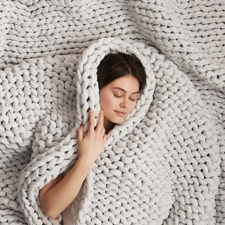 A woman with closed eyes is wrapped in a thick, white chunky knit blanket, surrounded by its textured folds.