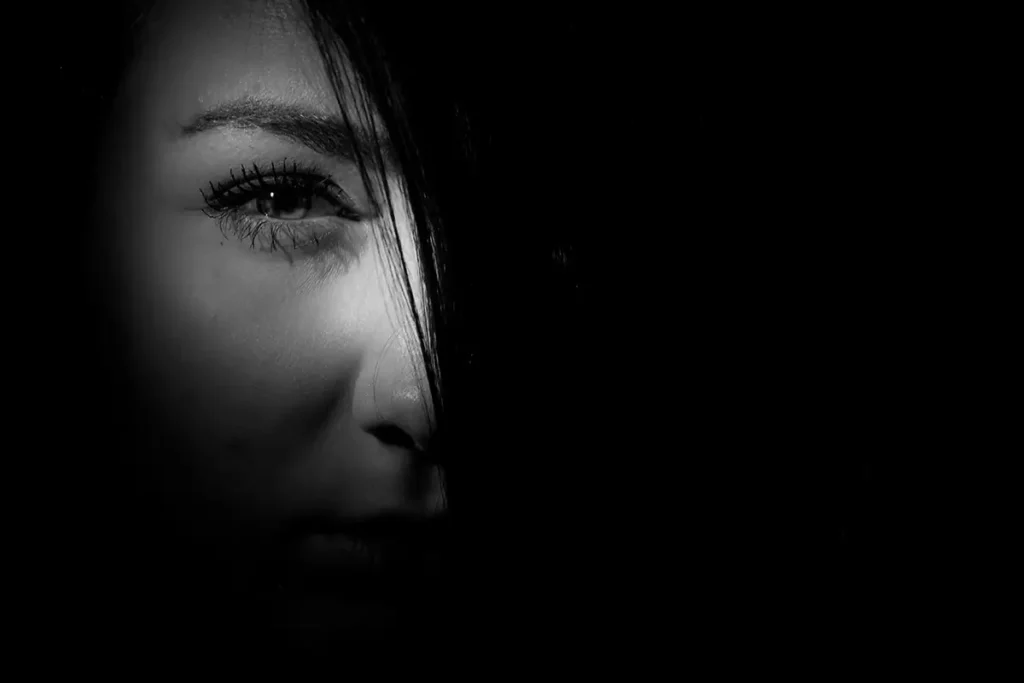 Black-and-white close-up of a woman’s face partially obscured by deep shadow, with one eye and part of her expression illuminated, creating a moody, dramatic contrast